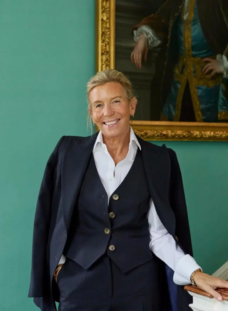 A sophisticated woman in a tailored navy waistcoat and blazer smiles elegantly against a vibrant teal wall with a classic portrait behind her.